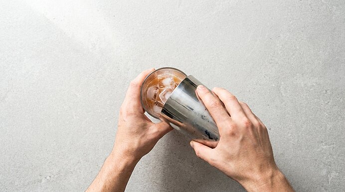 Zwei Hände halten einen Metall-Shaker über ein Glas mit Eis und schenken einen Cocktail ein.