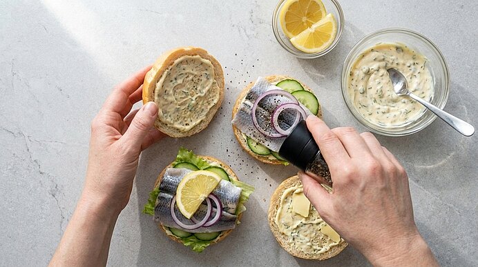 Eine Hand mahlt frisch Pfeffer über ein belegtes Fischbrötchen mit Bismarckhering, Gurke und Zwiebelringen.