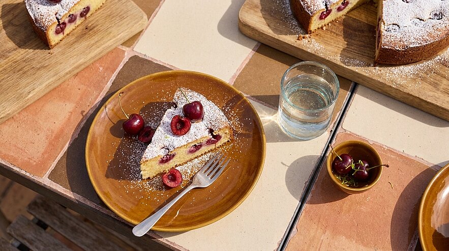 Eine aufgeschnittene Kirschkuchen-Scheibe auf braunem Teller, mit Puderzucker bestäubt und frischen Kirschen garniert; im Bild weitere Kuchenstücke auf Holzbrett und ein Glas Wasser.