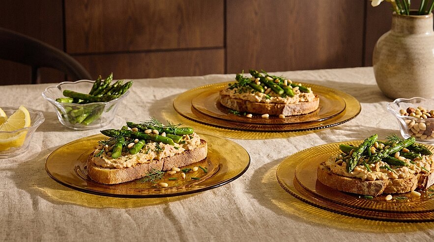 Drei offene Sandwiches auf Tellern: dicke Brotscheiben mit cremigem Aufstrich, grünen Spargelspitzen und Pinienkernen, angerichtet auf einem Leinentisch.