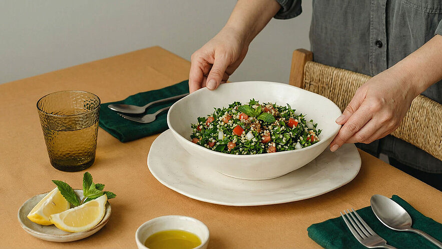 Eine Hand platziert eine große Schüssel mit Tabouleh, einem Salat aus Petersilie, Tomaten und Bulgur, auf einem Tisch. Neben der Schüssel liegt ein Teller mit Zitronenscheiben und frischer Minze. Im Hintergrund steht ein Getränk in einem Glas.