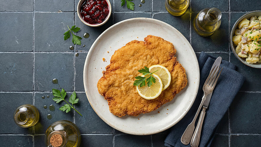Ein großflächiges, paniertes Schnitzel liegt auf einem weißen Teller, garniert mit zwei Zitronenscheiben und etwas Petersilie. Daneben befindet sich eine Schüssel mit Kartoffelsalat und eine kleine Schale mit Preiselbeeren. Der Hintergrund ist aus grauen Fliesen.
