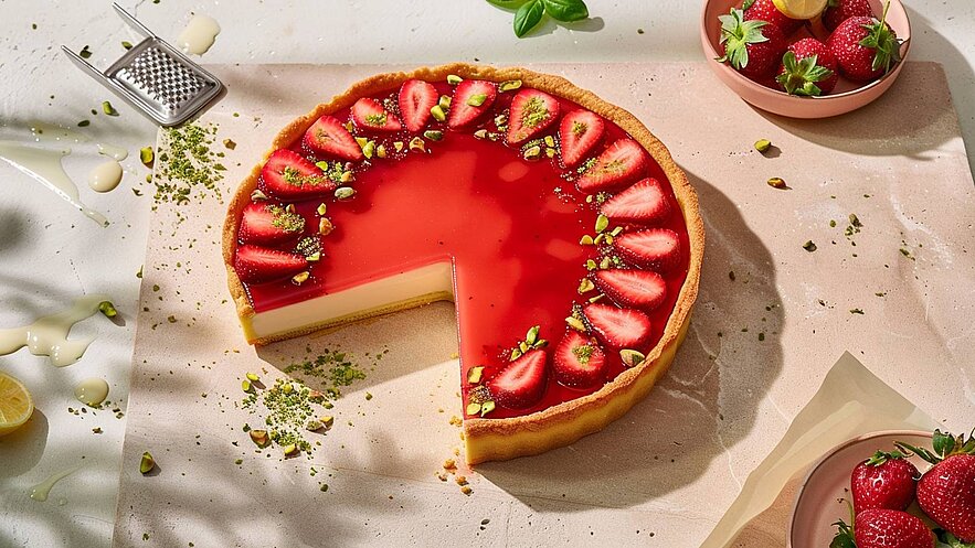 Ein Erdbeerkuchen mit einer glänzenden roten Glasierschicht und frischen Erdbeeren oberhalb. Der Kuchen hat einen goldbraunen Boden und ist teilweise angeschnitten. Um den Kuchen herum liegen gehackte Pistazien und einige Erdbeeren in einer kleinen Schale.