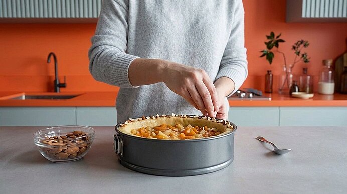 Hände einer Person bestreuen einen Obstkuchen in einer Springform mit gehackten Nüssen; Schüssel mit Mandeln und Löffel daneben.