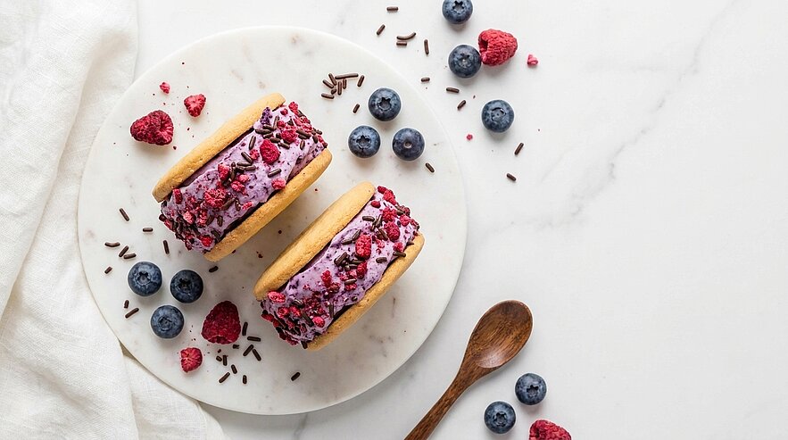 Zwei Eis-Sandwiches mit rosa-violettem Beereneis und zerkrümelten Beeren auf weißem Teller, garniert mit frischen Blaubeeren, Himbeeren und Schokostreuseln.