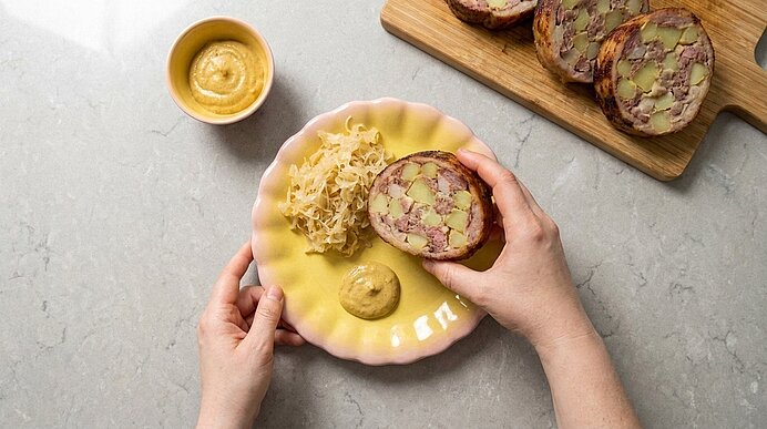 Hände halten eine Scheibe Wurstpastete über einem Teller mit Senf und Sauerkraut; auf einem Brett liegen mehrere weitere Scheiben und eine kleine Schale mit Senf.