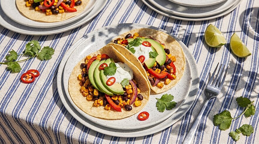 Zwei weiche Tacos auf einem Teller, gefüllt mit Mais, schwarzen Bohnen, roten Paprikastreifen, Avocadoscheiben, saurer Sahne, frischem Koriander und Chilischeiben; Limettenkeile daneben.