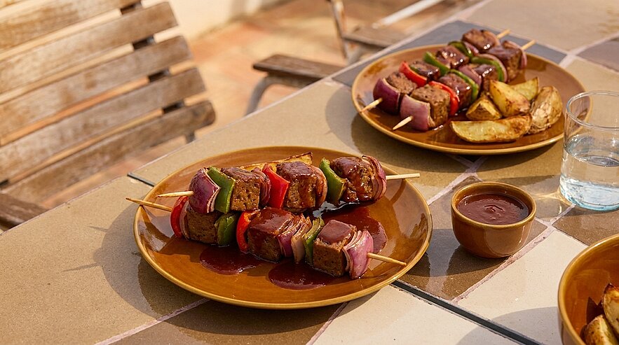 Zwei Teller mit gegrillten Fleisch- und Gemüsespießen (Paprika, Zucchini, Zwiebel), dazu gebackene Kartoffelspalten und eine Schale dunkler Barbecue‑Sauce.