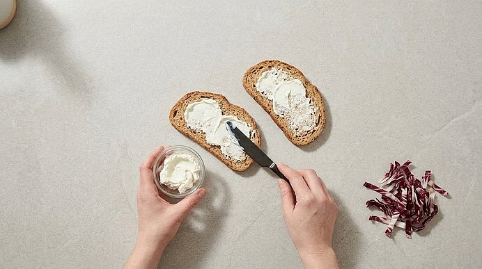 Zwei Hände bestreichen Vollkornbrotscheiben mit Ziegenfrischkäse; daneben liegen geschnittene Radicchiostreifen.