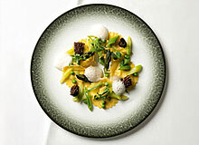 Auf dem Bild ist ein Teller mit frischen Ravioli zu sehen, garniert mit grünem Spargel, Morcheln und kleinen Schaumkugeln. Die Pastagerichte sind in einer ansprechenden Anordnung präsentiert und weisen eine leuchtend gelbe Farbe auf, eingerahmt von einem eleganten Rand.