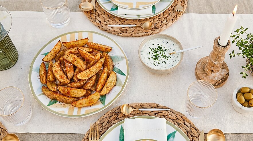 Teller mit würzigen Kartoffelspalten und Schälchen mit Kräuter-Dip auf gedecktem Esstisch, umgeben von Tellern, Gläsern, Besteck und einer brennenden Kerze.