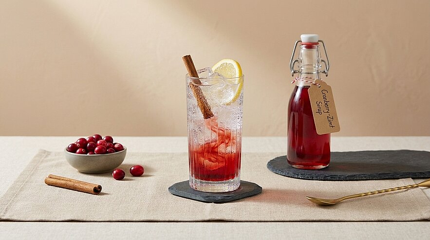 Hohes Glas mit Eis, rotem Cranberry-Drink, Zitronenscheibe und Zimtstange neben einer Flasche hausgemachtem Cranberry-Sirup und einer kleinen Schale frischer Cranberries auf hellem Tisch.
