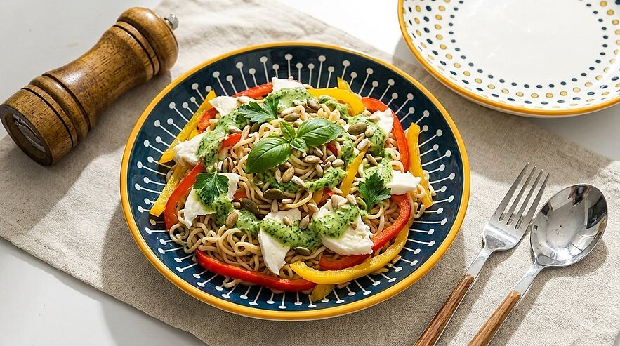 Bunter Nudelsalat auf blauem Teller mit gelben und roten Paprikastreifen, Mozzarellastücken, grünem Pesto, frischem Basilikum und gerösteten Kernen.