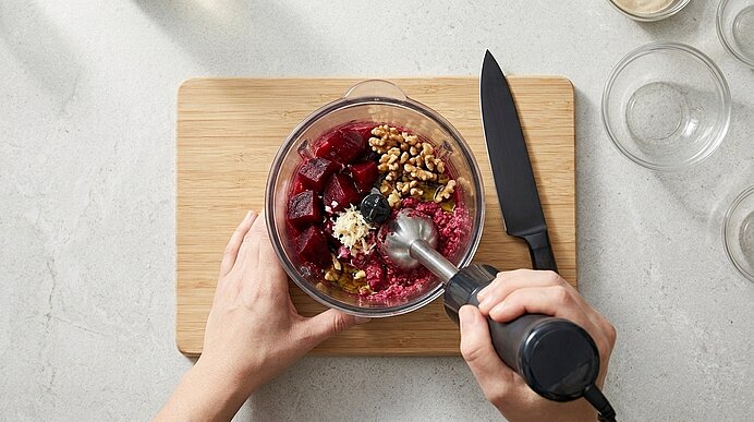 Zwei Hände pürieren Rote Rüben, Walnüsse und Kren in einem Becher mit einem Stabmixer auf einem Holzbrett.