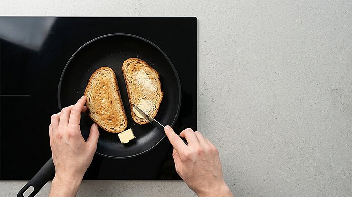 Zwei Hände bestreichen geröstete Brotscheiben in einer Pfanne auf dem Kochfeld mit Butter.