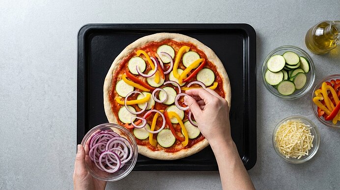 Hände legen rote Zwiebelringe auf den mit Tomatensauce, Zucchini und Paprika belegten Pizzateig auf einem Backblech.