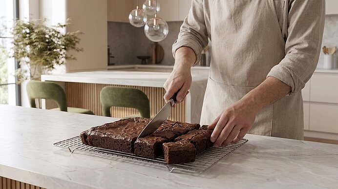 Person in Schürze schneidet mit großem Messer frisch gebackene Schokoladen-Brownies auf einem Abkühlgitter in der Küche in gleichmäßige Quadrate.