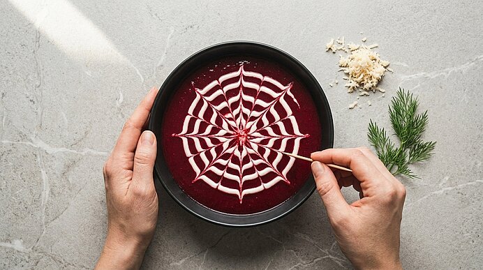 Zwei Hände ziehen mit Zahnstocher ein Sauerrahm-Spinnennetz auf Rote-Bete-Suppe; daneben liegen Meerrettich und Dill.