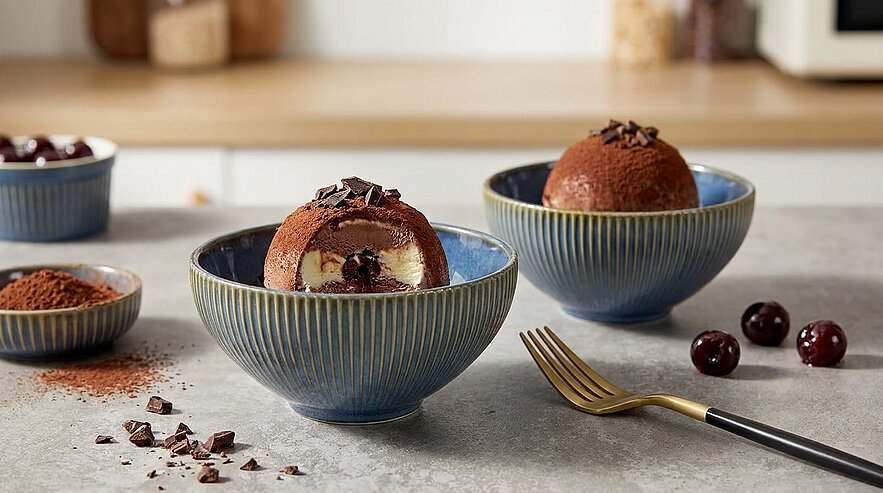 Zwei blaue Schalen mit schokoladenüberzogenen Dessertkuppeln; eine halb aufgeschnitten, zeigt Cremefüllung und Kirschfüllung, garniert mit Schokoraspeln, daneben frische Kirschen und eine Gabel.