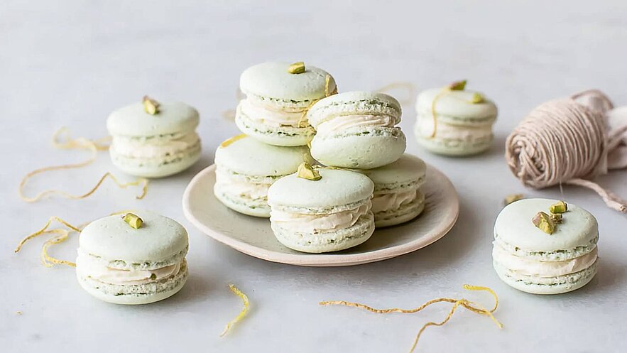 Ein Teller mit mehreren grünen Macarons, gefüllt mit einer hellen Creme, ist auf einem hellen Untergrund platziert. Einige Macarons liegen daneben, und einige sind mit gehackten Pistazien dekoriert. Die Anordnung ist ansprechend und sättigend.