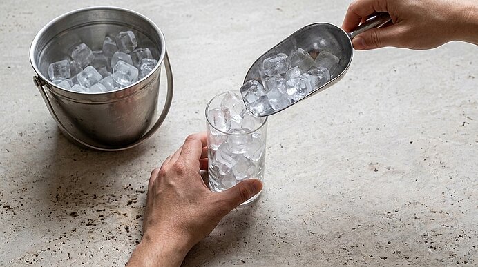 Hände füllen ein hohes Glas mit Eiswürfeln aus einer Metallschaufel; daneben ein Metall-Eiseimer.