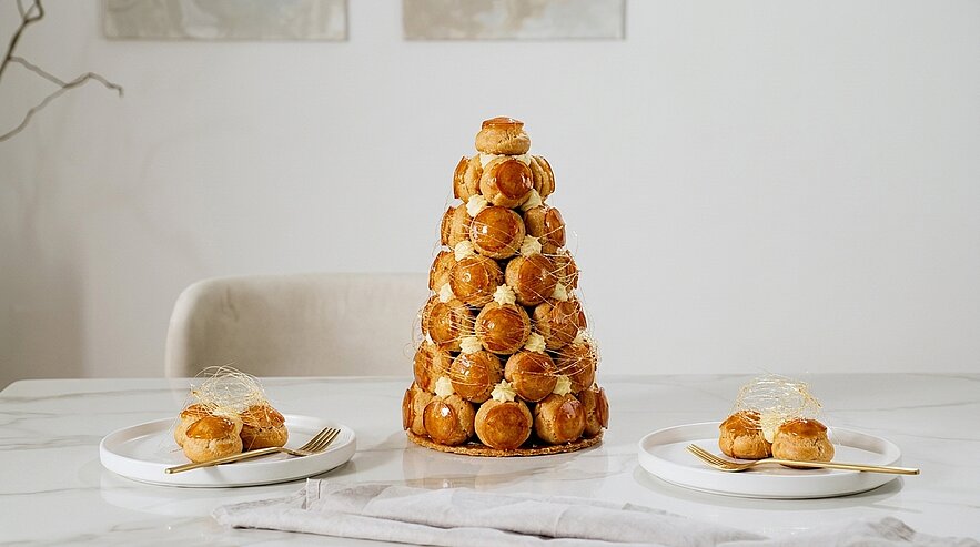 Hohe, kegelförmige Croquembouche aus Windbeuteln mit Karamellspinnweben, auf einem Esstisch, daneben zwei Teller mit je drei Windbeuteln und Besteck.