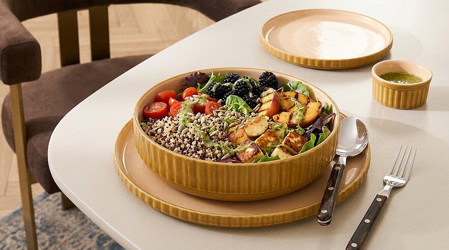 Bunte Schüssel mit Buddha-Bowl: Blattsalat, Quinoa, gegrillte Käsewürfel, Kirschtomaten, Brombeeren und Nüsse; Besteck daneben.