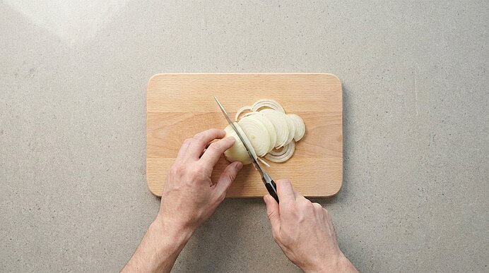 Zwei Hände schneiden eine weiße Zwiebel in gleichmäßige Scheiben mit einem Küchenmesser auf einem Holzschneidebrett.