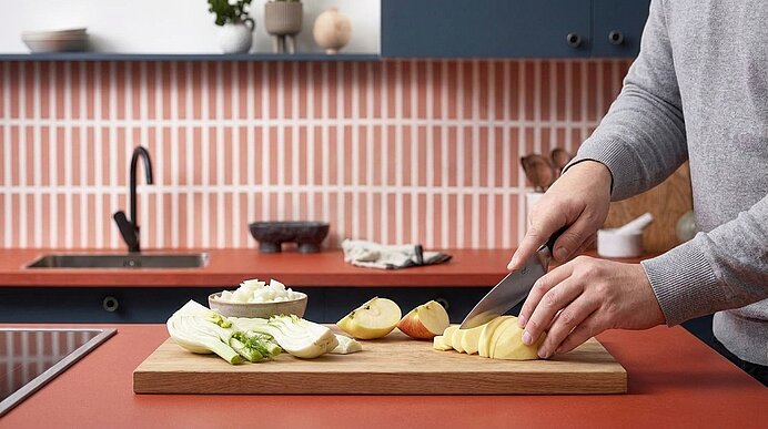 Auf einem Holzbrett schneidet eine Person Kartoffelscheiben in einer modernen Küche; daneben liegen Fenchel, Spargel und eine Schale mit gehackten Zwiebeln.