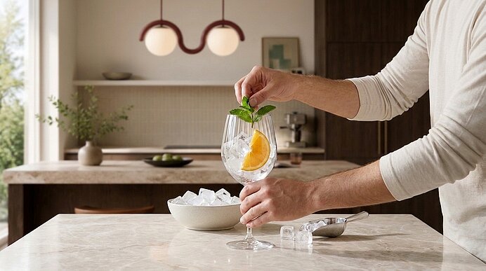Hände platzieren frische Minze und eine Orangenscheibe in ein Weinglas mit Eis auf einer Küchenarbeitsplatte bei der Cocktailzubereitung.