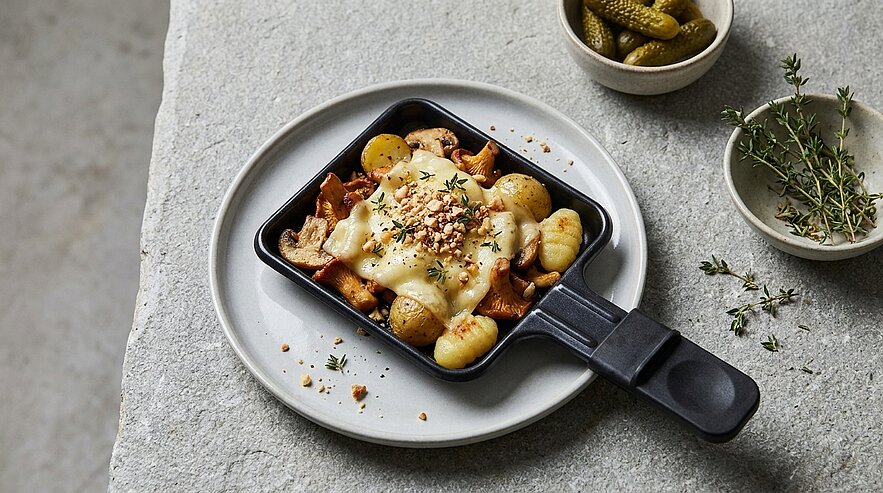 Kleines Raclette-Pfännchen mit geschmolzenem Käse über Kartoffelgnocchi und Pilzen, garniert mit gehackten Nüssen und frischen Kräutern auf einem weißen Teller.