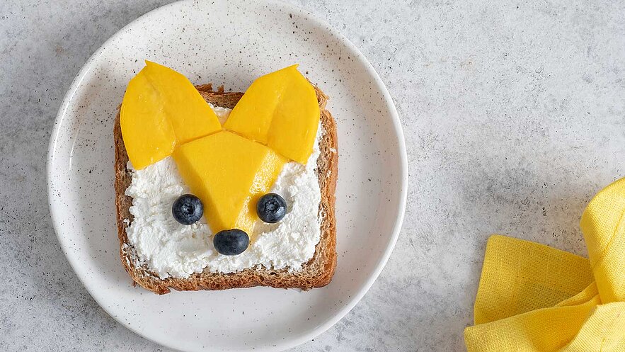Ein Stück Vollkornbrot liegt auf einem Teller. Darauf ist eine kreative Gestaltung mit Frischkäse, Mango und Heidelbeeren, die ein Tiergesicht darstellen. Die Ohren sind aus Mangoscheiben, und die Augen sowie die Nase sind aus Heidelbeeren.
