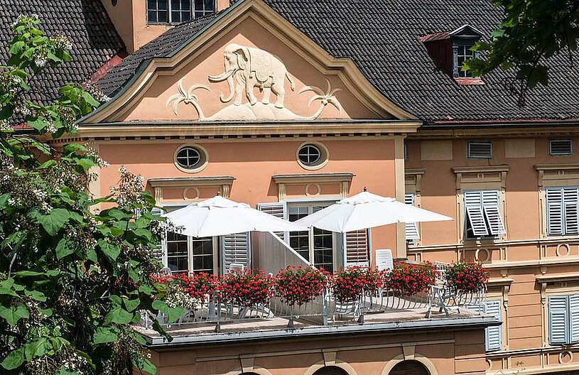 Balkonterrasse eines historischen, hellorangefarbenen Gebäudes mit weißen Sonnenschirmen, roten Balkonblumen, dekorativem Fassadenrelief und geschlossenen Fensterläden.