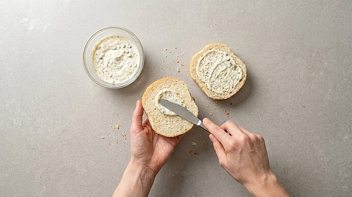 Zwei Hände bestreichen eine Brotscheibe mit Kräuterfrischkäse; daneben liegt eine bereits bestrichene Scheibe und ein Glas mit cremigem Aufstrich.