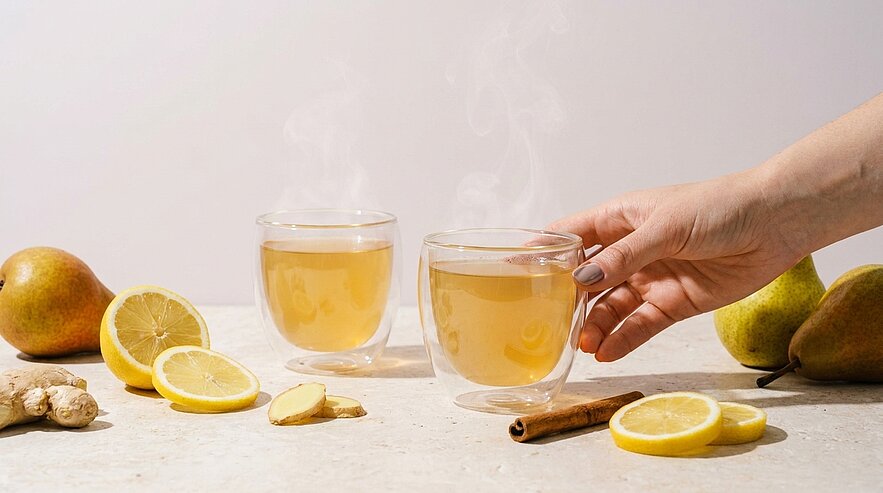 Zwei dampfende Glastassen mit heißem Ingwer‑Zitronen‑Tee; eine Hand greift nach einer Tasse. Vorne Zitronenscheiben, Ingwerstücke und eine Zimtstange, im Hintergrund Birnen.