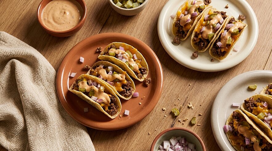 Drei knusprige Maistacos gefüllt mit gewürztem Hackfleisch, roten Zwiebelwürfeln und cremiger Sauce, auf Tellern angerichtet mit kleinen Schälchen Beilagen.