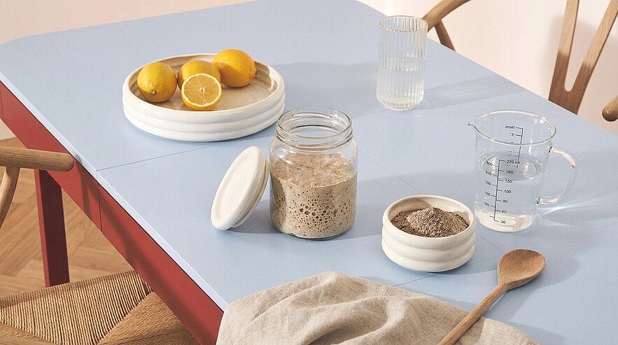 Küchentisch mit offenem Glasgefäß aktivem Sauerteig, kleine Schale mit Mehl, Messbecher mit Wasser, Teller mit Zitronen, Trinkglas, Holzlöffel und Küchentuch.