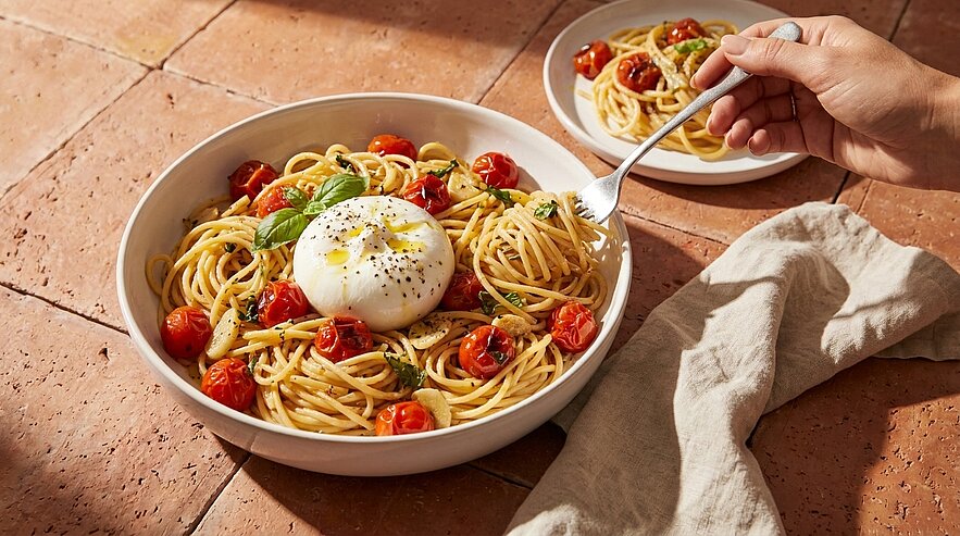 Schüssel Spaghetti mit Kirschtomaten, frischem Basilikum und einer Burrata‑Kugel in der Mitte; daneben eine Hand, die mit einer Gabel etwas Pasta aufnimmt.