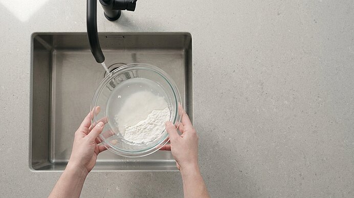 Zwei Hände halten eine Glasschüssel mit Mehl und Wasser über einer Edelstahl-Küchenspüle, im Bild ein schwarzer Wasserhahn zu sehen.