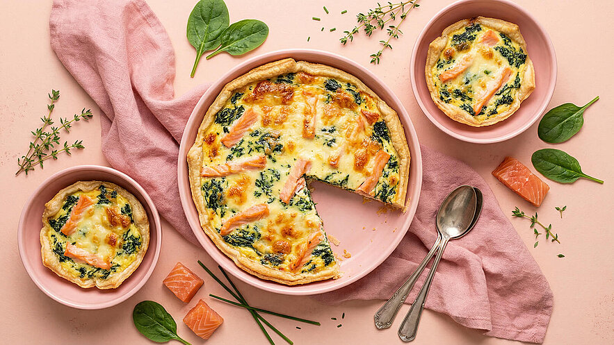 Auf dem Bild ist eine große Quiche mit Lachs und Spinat zu sehen, aus der ein Stück herausgeschnitten wurde. Daneben stehen zwei kleine Portionen in rosa Schalen. Frische Kräuter und einige Stücke Lachs ergänzen das Bild. Der Hintergrund ist in sanften Pastellfarben gehalten.