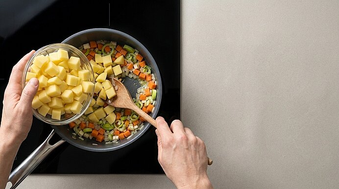Hände geben gewürfelte Kartoffeln aus einer Schüssel in eine Pfanne mit angebratenem Sellerie, Karotten und Zwiebeln, während mit einem Holzlöffel gerührt wird.