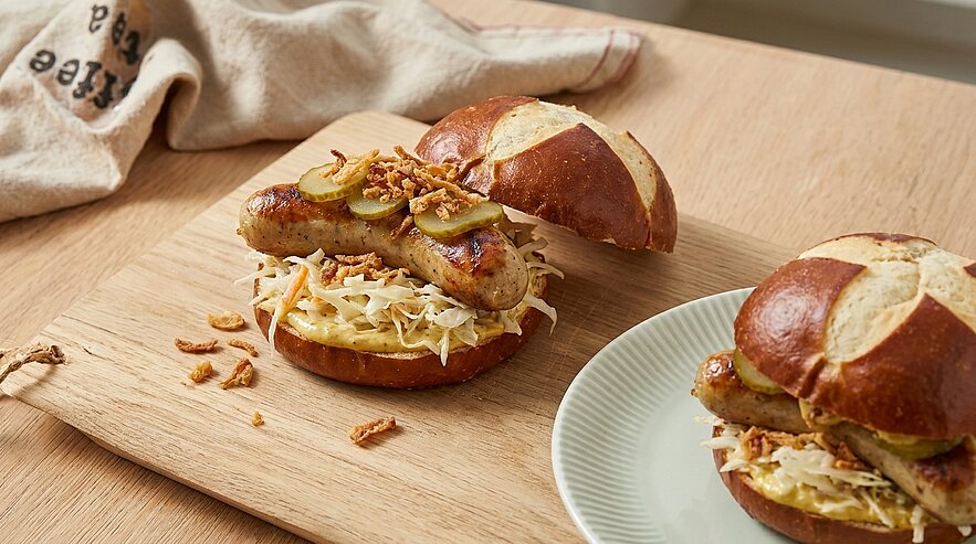 Zwei Bratwürste in Laugenbrötchen, belegt mit Sauerkraut/Krautsalat, Senf, Gewürzgurken und knusprigen Röstzwiebeln auf einem Holzbrett.