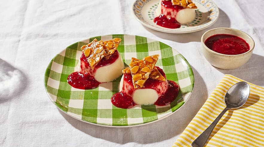 Zwei Panna-Cotta-Portionen mit roter Beerensauce und knusprigem Mandelkrokant auf einem grünen karierten Teller.