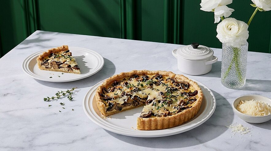 Herzhafte Pilz-Quiche auf weißem Teller, ein Stück herausgeschnitten und auf einem separaten Teller serviert, mit geschmolzenem Käse und frischem Thymian garniert.