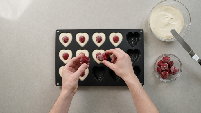 Zwei Hände legen gefrorene Himbeeren in herzförmige Backförmchen mit hellem Käsekuchenteig; daneben eine Schüssel mit Teig und eine Schale mit Himbeeren.