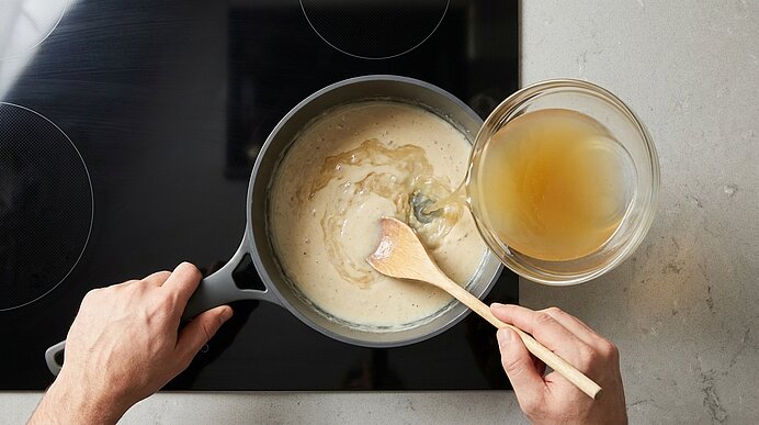 Zwei Hände rühren mit einem Holzlöffel eine cremige helle Sauce in einem Topf auf dem Herd; daneben steht eine Glasschüssel mit Brühe zum Unterrühren.