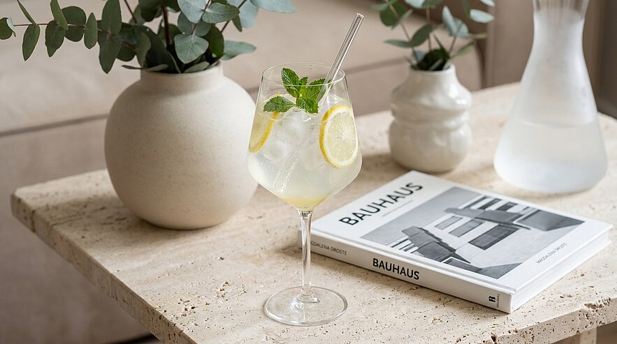 Weinglas mit eisgekühltem Zitronen-Minz-Getränk und Strohhalm auf hellem Steintisch neben einem Buch mit dem Titel "Bauhaus" und zwei dekorativen Vasen.