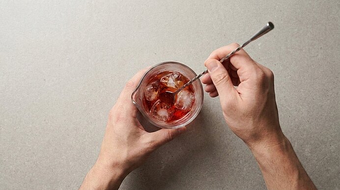 Von oben: Zwei Hände halten ein kurzes Glas mit Eiswürfeln; ein langer Löffel rührt ein rötlich-braunes Getränk.