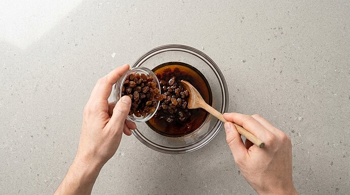 Hände geben Rosinen aus einer kleinen Schüssel in eine Glasschüssel mit dunkler Flüssigkeit und rühren mit einem Holzlöffel.