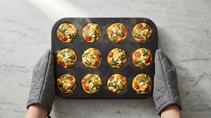 Blech mit zwölf frisch gebackenen Mini‑Quiches/Ei‑Muffins mit Gemüsefüllung, gehalten von zwei Händen in grauen Ofenhandschuhen.